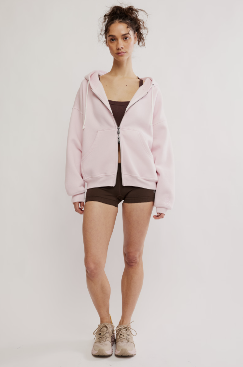 A woman modeling a light pink zip-up hoodie over a dark brown sports bra and matching shorts, paired with beige sneakers, against a plain light gray studio background.