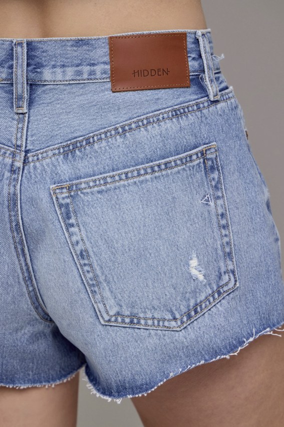 Close-up of light blue denim shorts with a brown patch label on a neutral background