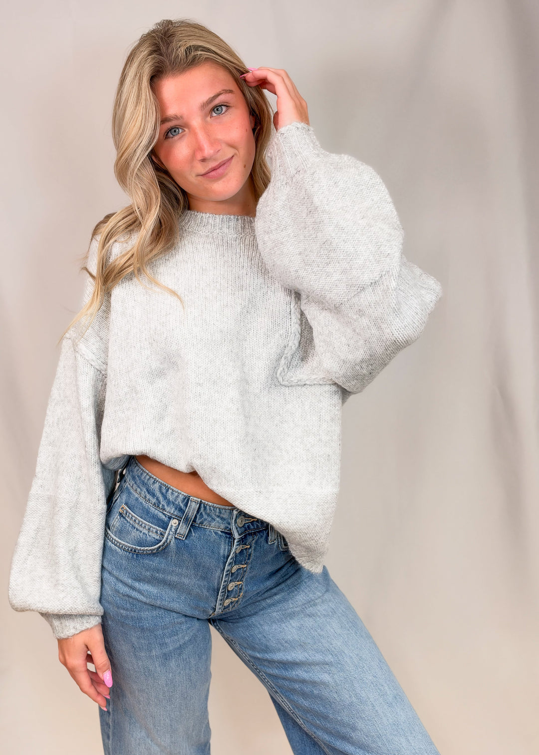 A woman modeling a light gray fuzzy cropped sweater with voluminous balloon sleeves, paired with blue jeans, against a neutral beige studio backdrop.
