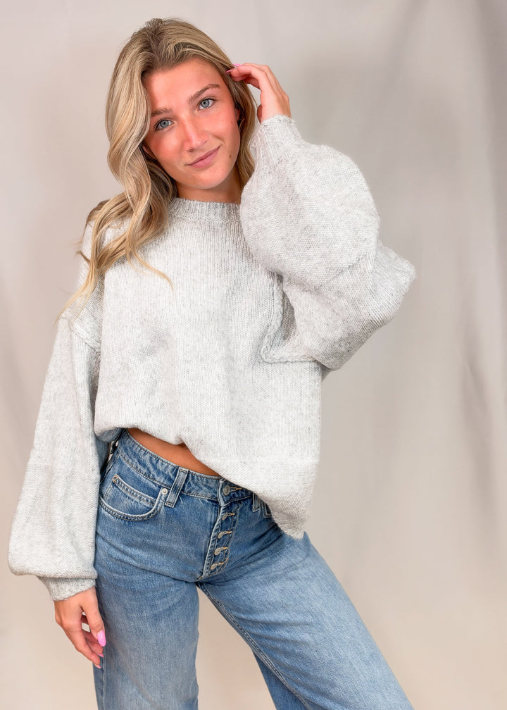A woman modeling a light gray fuzzy cropped sweater with voluminous balloon sleeves, paired with blue jeans, against a neutral beige studio backdrop.