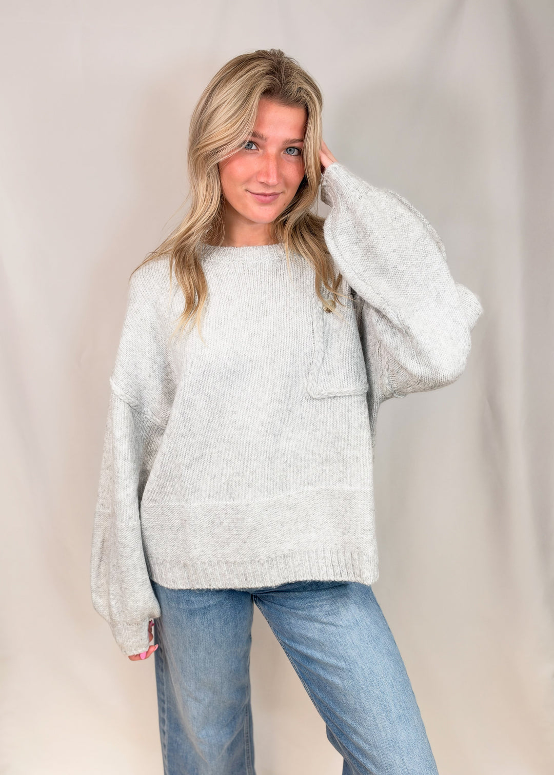 A woman modeling a light gray heathered knit sweater with voluminous balloon sleeves and a front chest pocket, paired with blue jeans, against a neutral draped backdrop.