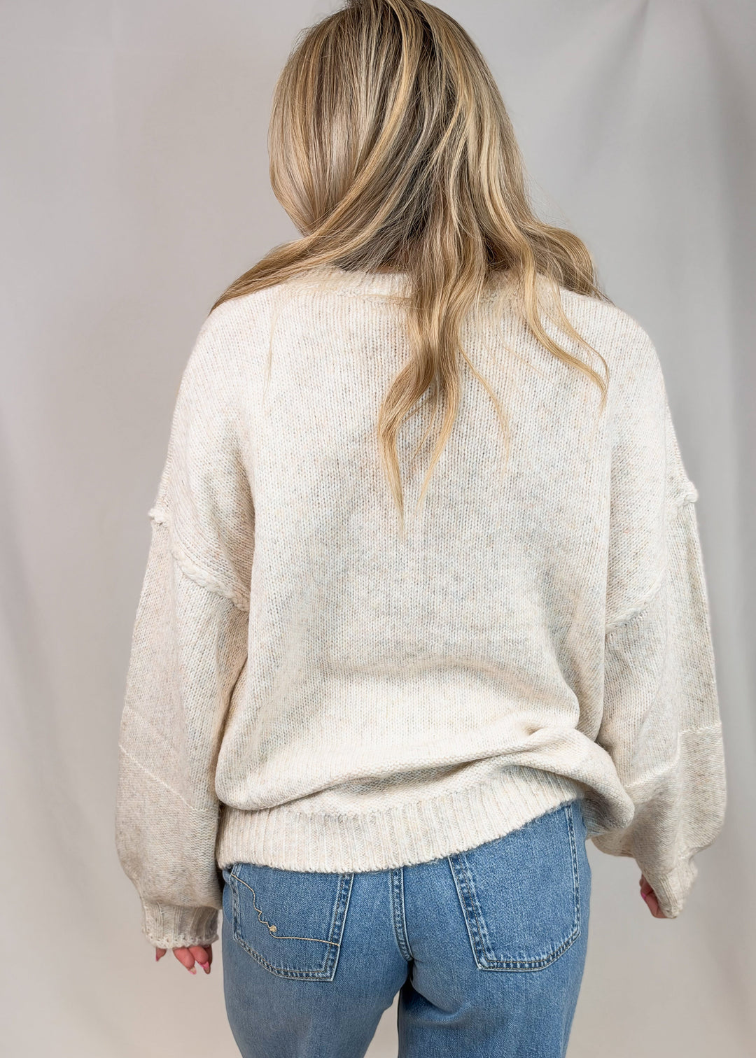 A woman viewed from behind wearing a cream-colored, textured knit sweater and blue jeans against a neutral studio backdrop.
