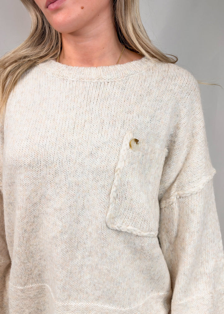 A close-up of a woman wearing a chunky knit sweater in a warm off-white or cream color. The sweater features a small decorative button detail on the chest pocket and a soft, fuzzy texture.