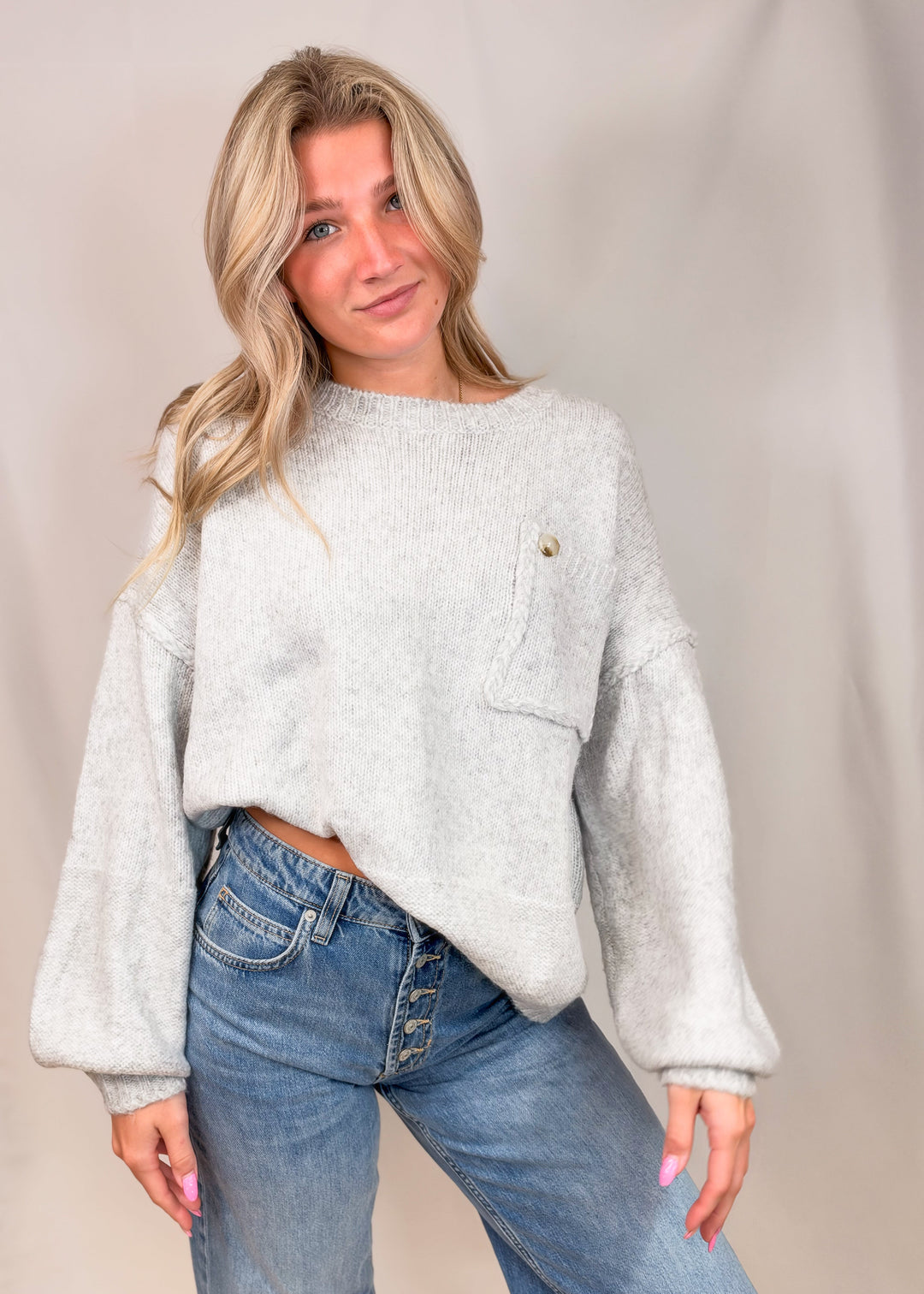 A woman wearing a light gray fuzzy knit sweater with a small gold button detail and blue jeans, standing against a neutral beige studio backdrop.