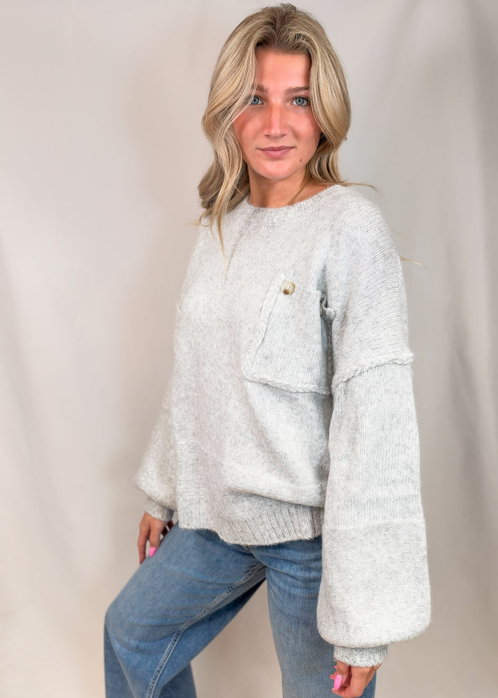 A woman wearing a light gray fuzzy knit sweater with a chest pocket and button detail, paired with blue jeans, posing against a neutral draped backdrop.