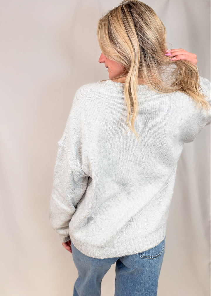 A woman with blonde hair is shown from behind wearing a light gray fuzzy knit sweater and blue jeans, posing against a neutral beige studio backdrop.