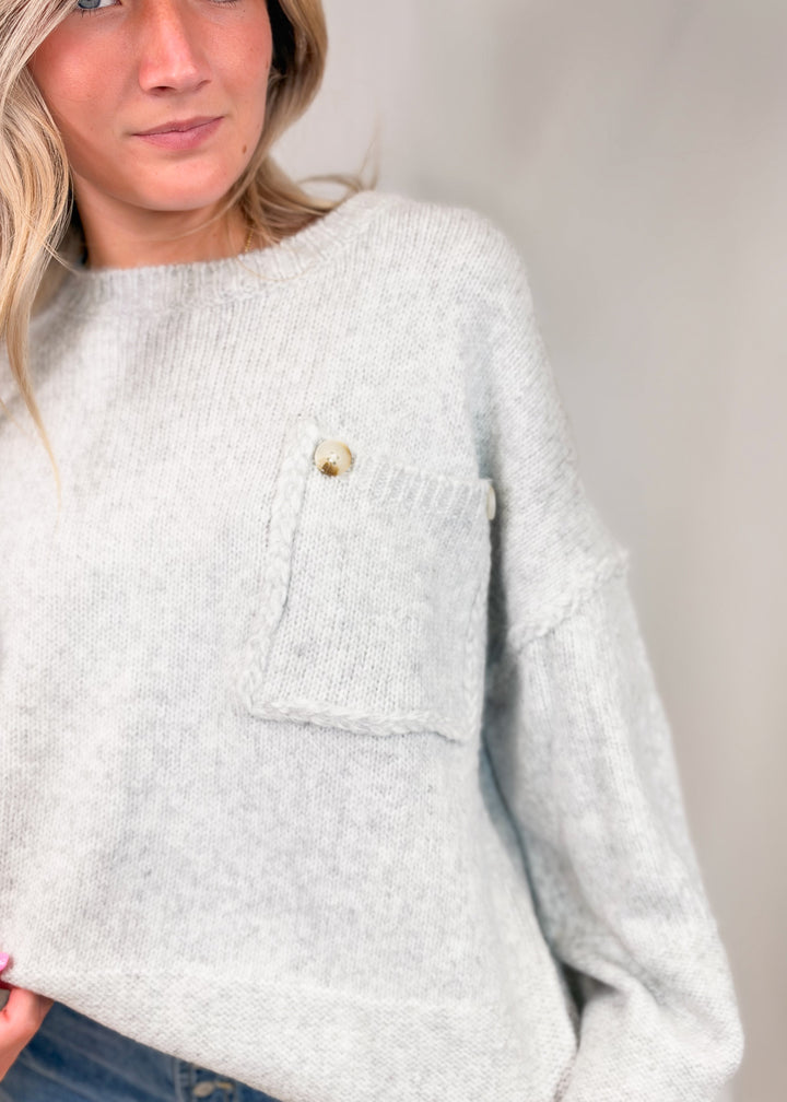 A close-up of a woman wearing a light heather gray knit sweater with a single gold button on the chest pocket, paired with blue jeans.