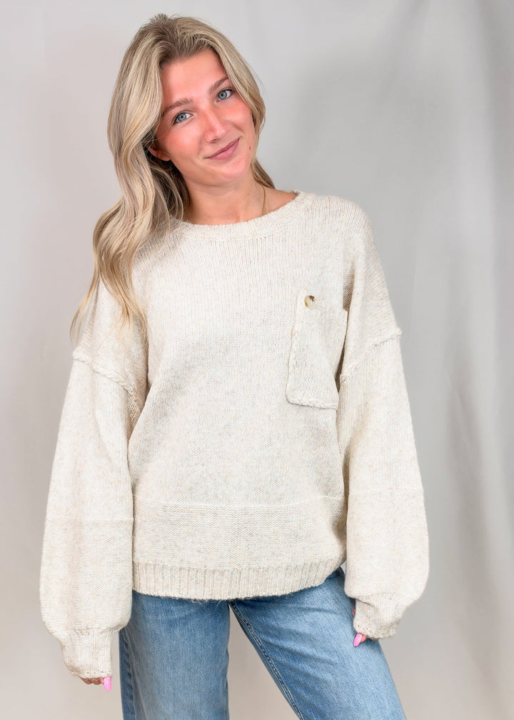 A woman wearing a cream-colored, textured knit sweater with a chest pocket and blue jeans, standing against a light gray studio backdrop.
