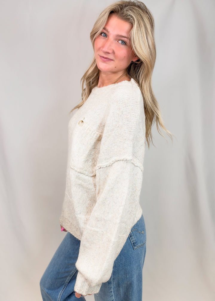 A woman with blonde hair wearing a cream-colored, textured knit sweater and blue jeans, posing against a light gray studio backdrop.