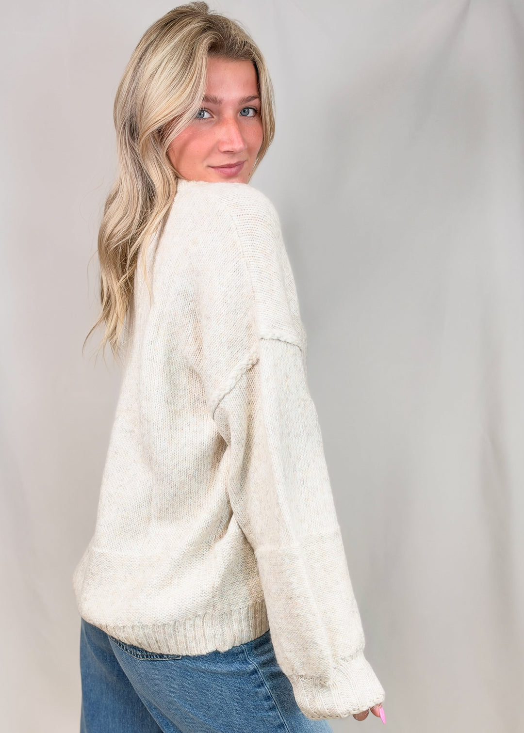 A woman wearing a fuzzy off-white knit sweater and blue jeans, photographed against a light neutral studio backdrop.