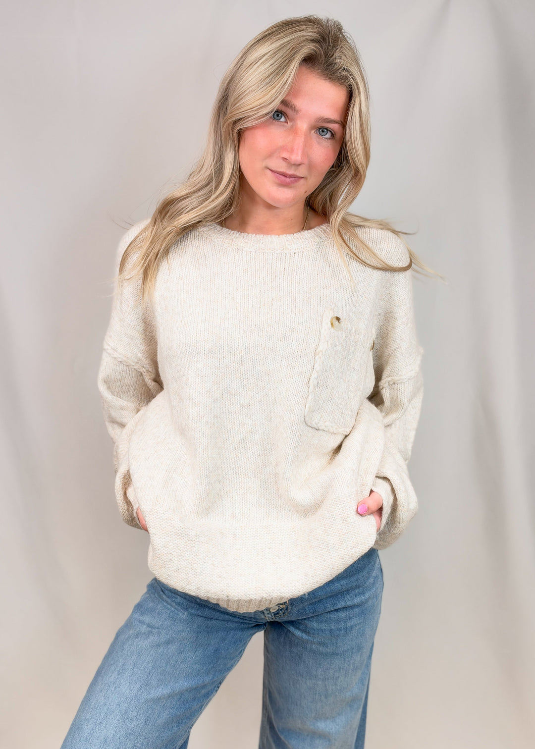 A woman modeling a cream-colored, textured knit sweater with a chest pocket and a small gold button, paired with blue jeans, against a neutral studio backdrop.