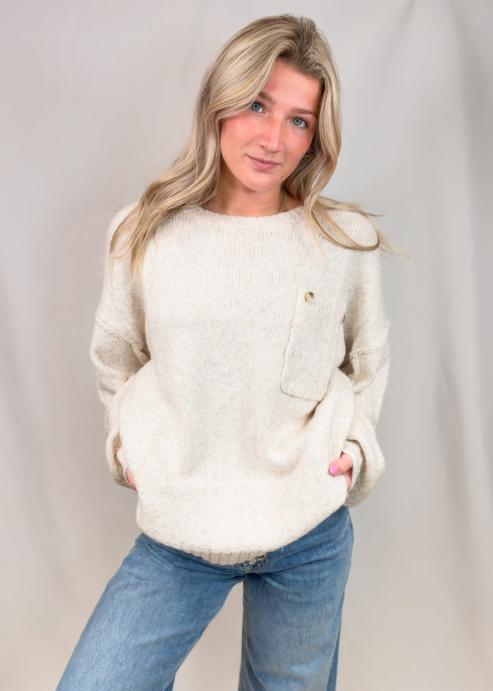 A woman modeling a cream-colored, textured knit sweater with a chest pocket and a small gold button, paired with blue jeans, against a neutral studio backdrop.