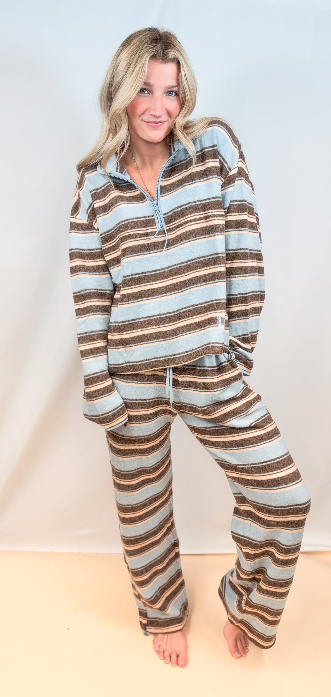 Woman wearing matching light blue and brown stripe set with quarter zip top and pants on plain background