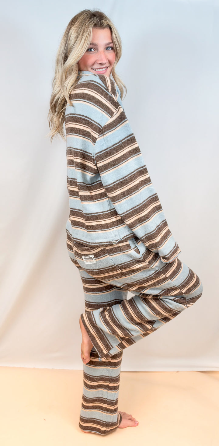 Woman shown from side view wearing matching light blue and brown stripe set with long sleeve top and pants on plain background