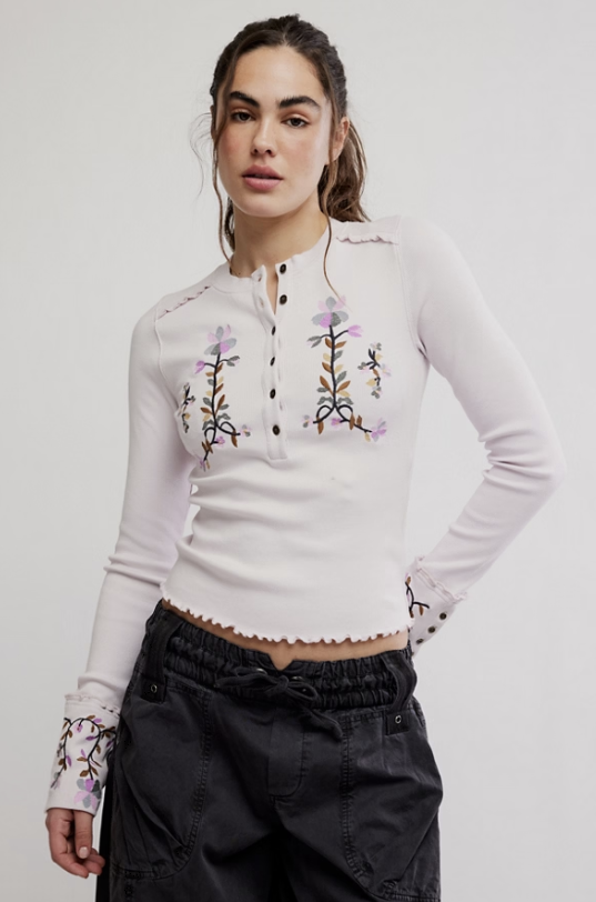 Woman wearing a white long-sleeve top with floral embroidery on a plain background