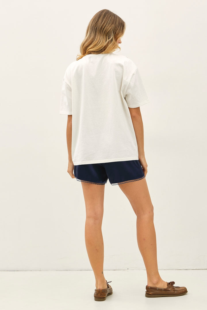 A woman shown from the back wearing an oversized white short-sleeve t-shirt, navy blue shorts with a thin white piping trim, and brown boat shoes against a plain white studio background.