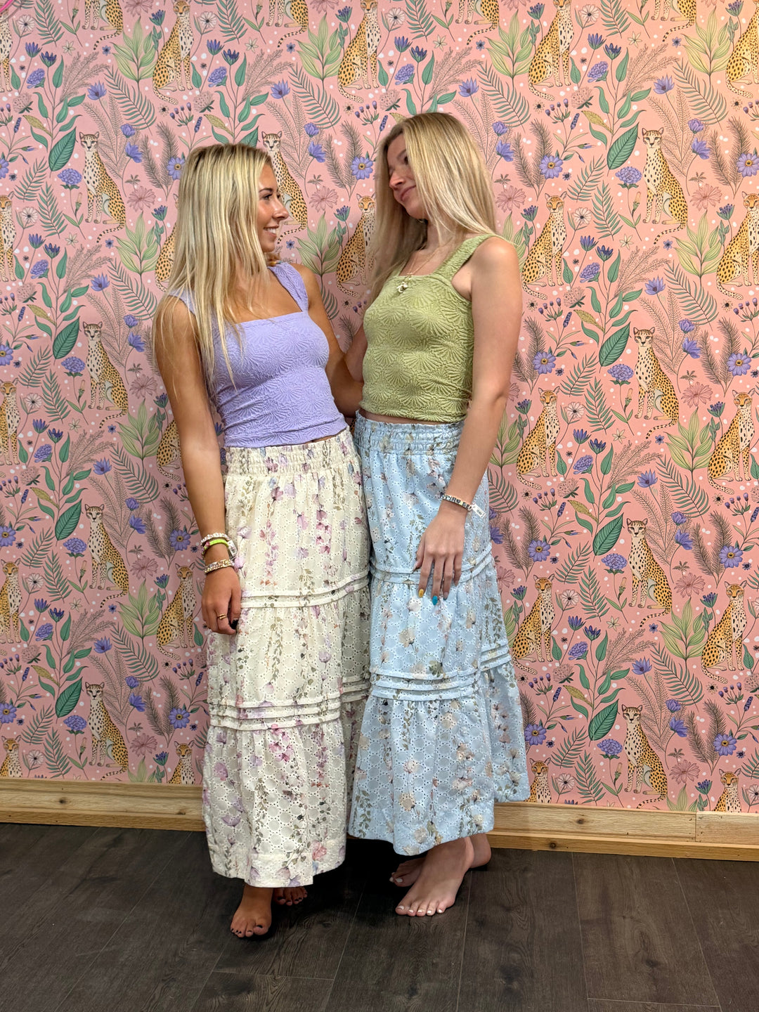 Two women standing against a pink floral-patterned wall.