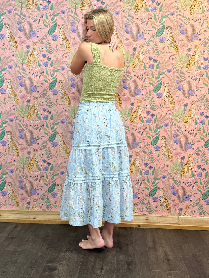 Woman wearing a green top and light blue floral skirt against a pink wallpapered wall with animal and floral patterns.