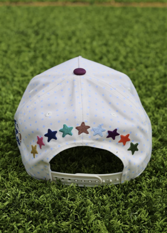 A white snapback cap with light blue polka dots and colorful embroidered star patches, photographed from the back on a green grass background.