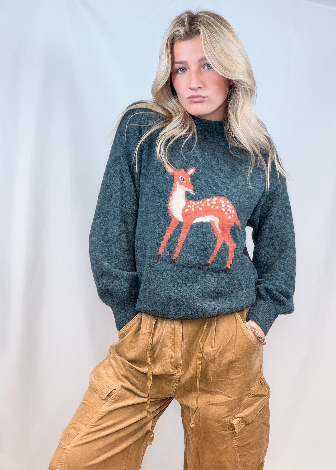 Woman wearing green sweater with fawn graphic on front with corduroy pants on plain background