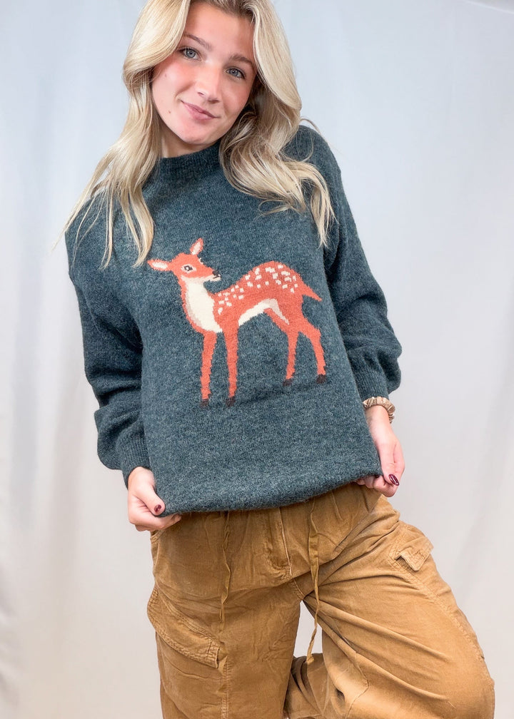 Woman wearing green sweater with fawn graphic and corduroy pants on plain background