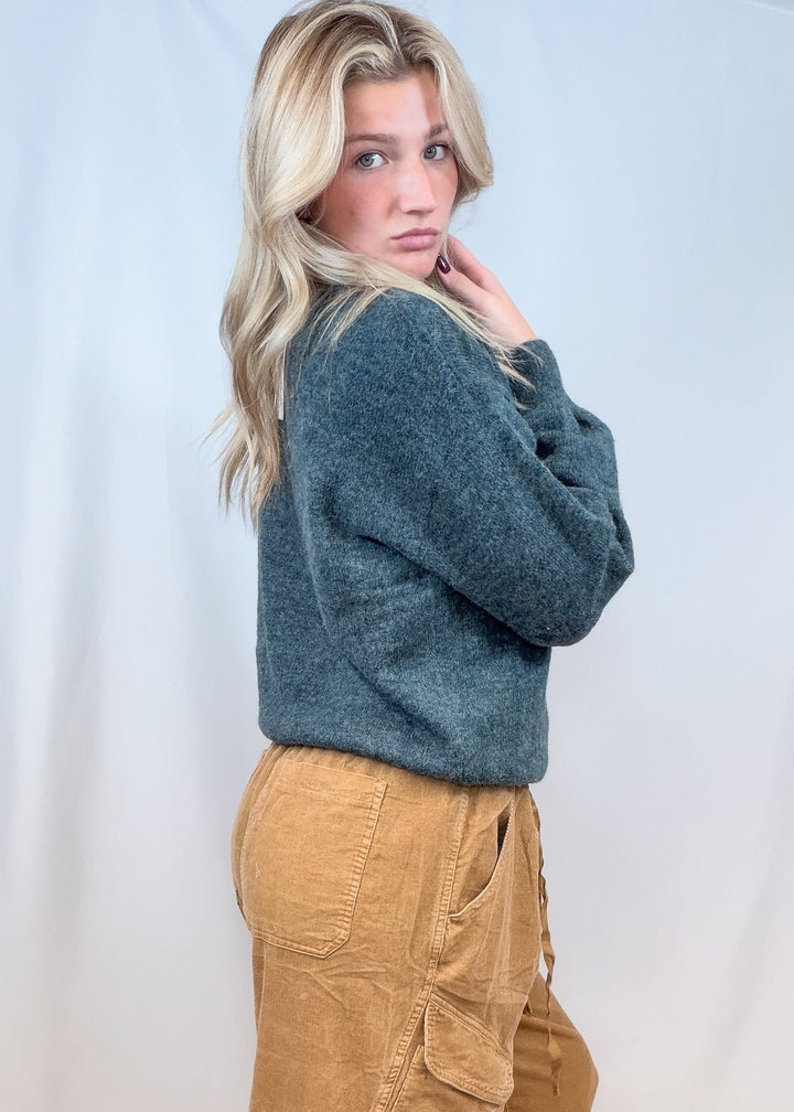 Woman shown from side view wearing green sweater and corduroy pants on plain background