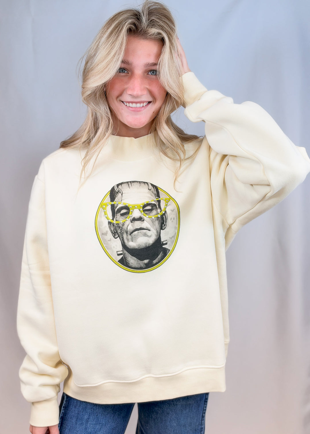 Woman wearing cream color mockneck with frankenstein graphic on front in yellow on plain background