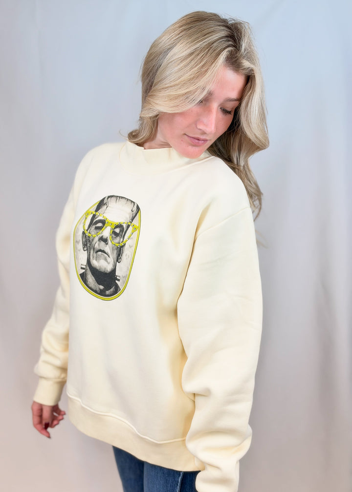 Woman wearing cream color mockneck with frankenstien graphic on front on plain background