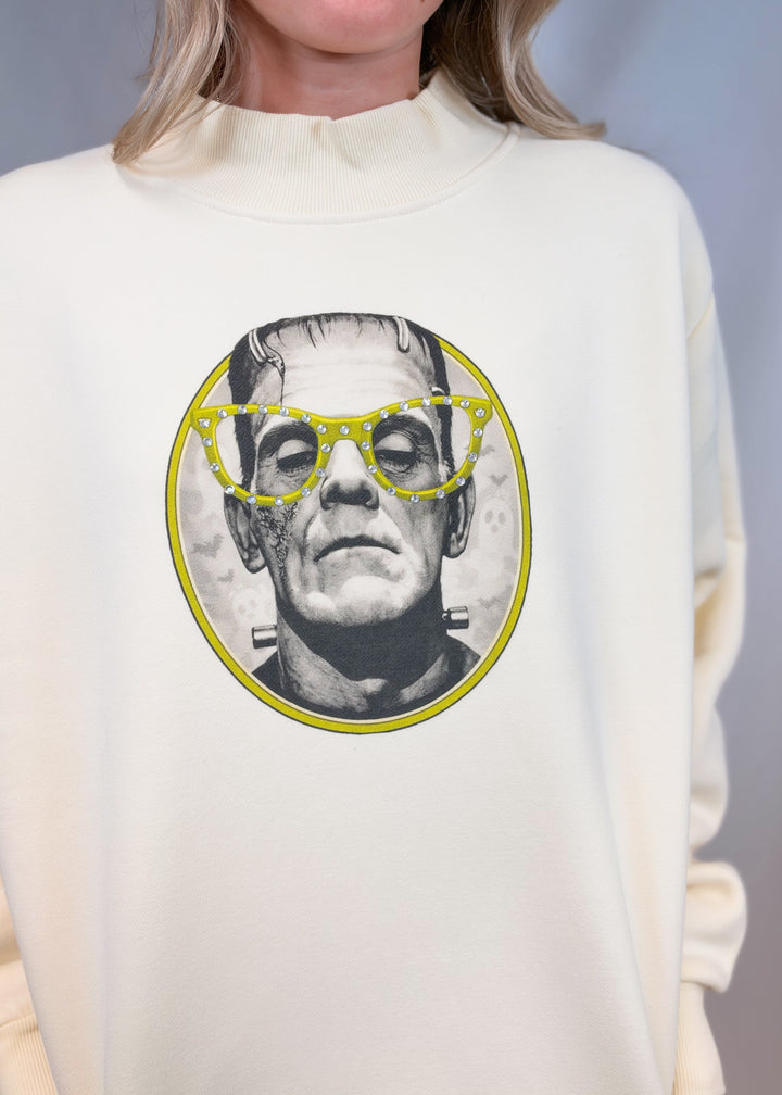 Close up of cream color mockneck with frankenstien graphic on front with yellow details and rhinestones on plain background