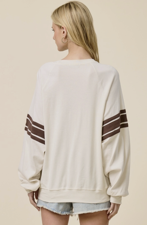 A woman shown from the back wearing an off-white crewneck sweatshirt with brown and white stripes on the sleeves, paired with denim shorts, against a solid beige background.