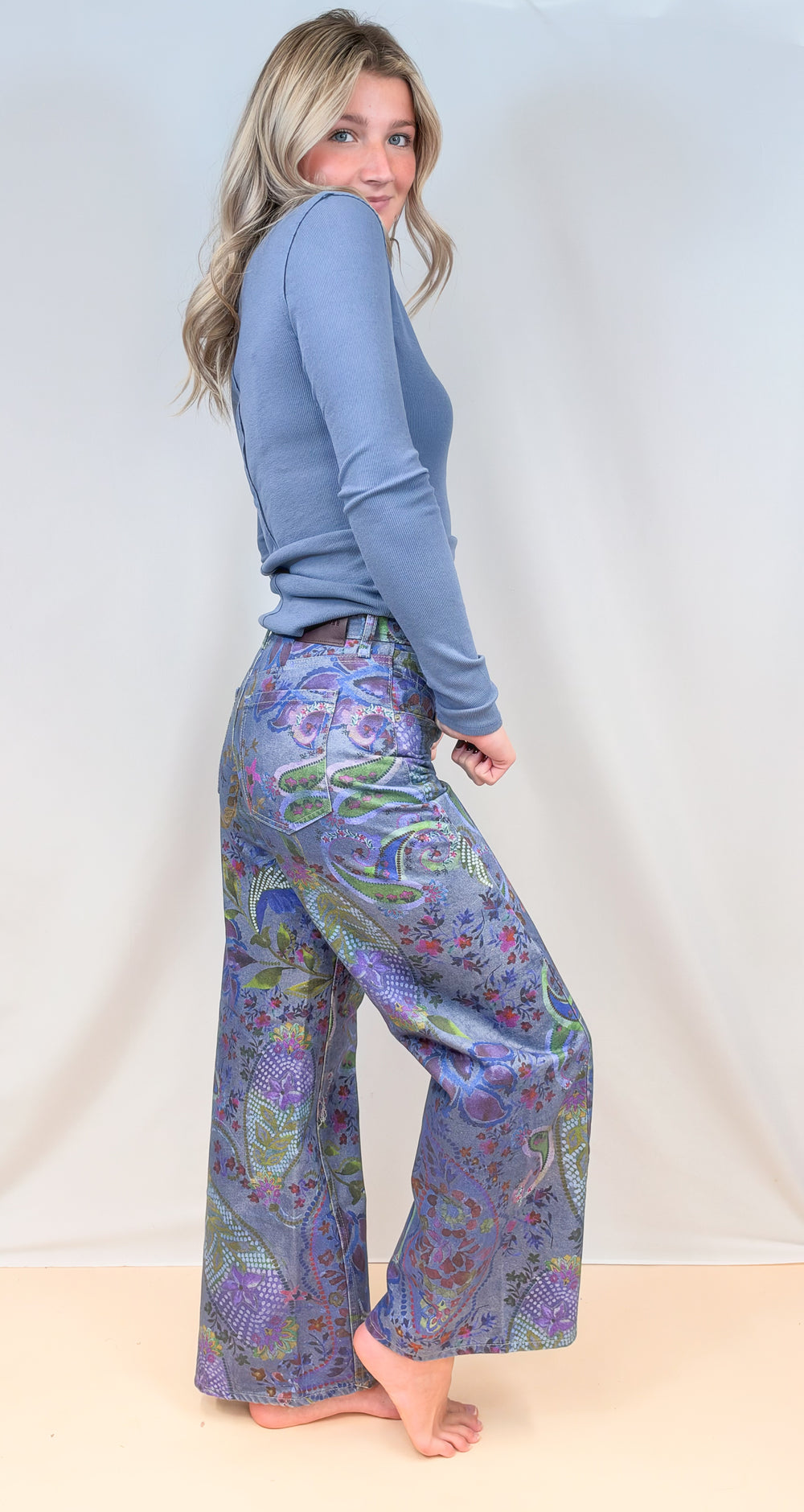 Woman shown from side view wearing blue paisley pants with blue long sleeve top on plain background