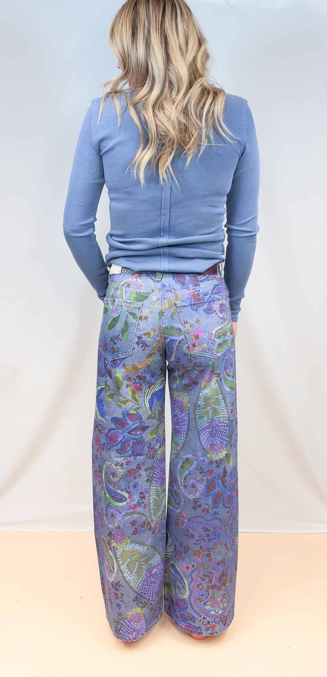 Woman shown from back view wearing blue paisley wide leg pants with blue long sleeve top on plain background
