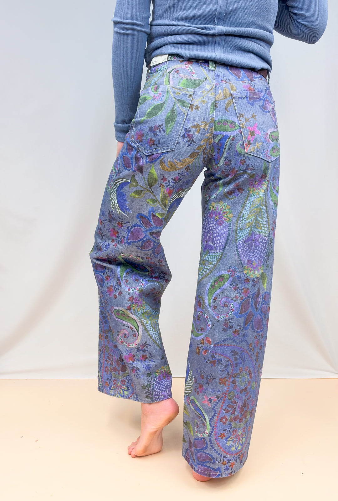 Woman shown from back view waist down wearing blue paisley jeans on plain background