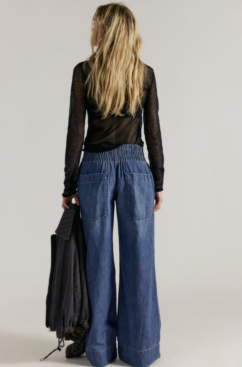 Woman shown from back wearing black long sleeve with medium wash wide leg denim jeans holding black jacket on plain background