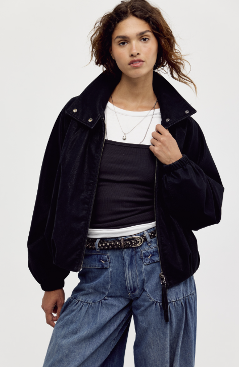 Woman wearing black suede jacket with white and black shirt under and jeans on plain background