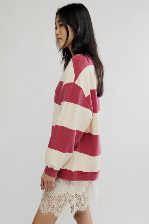 A woman shown from the side wearing an oversized long-sleeve knit sweater with wide horizontal stripes in deep pink and cream, paired with a white lace hem dress or skirt, against a plain light gray studio background.