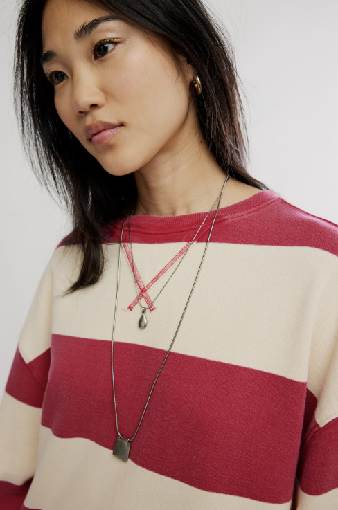 A close-up of a woman wearing a cream and red striped crewneck sweater, layered with two silver chain necklaces, against a plain light gray background.
