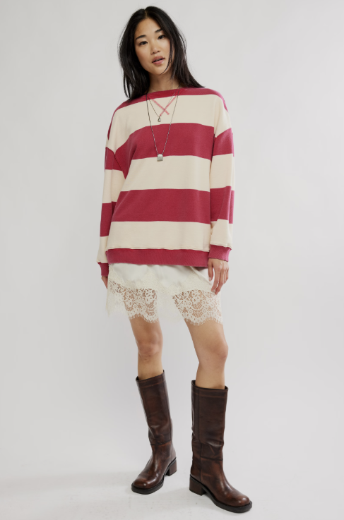 A model stands against a light gray studio background wearing a cream and red striped long-sleeve knit top, a white lace-trimmed mini skirt, and dark brown knee-high leather boots.