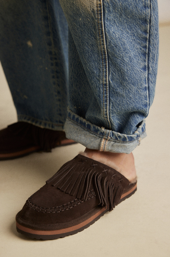 A close-up of a person wearing dark brown suede fringe mule slippers with rolled-up blue jeans, standing on a light beige floor.