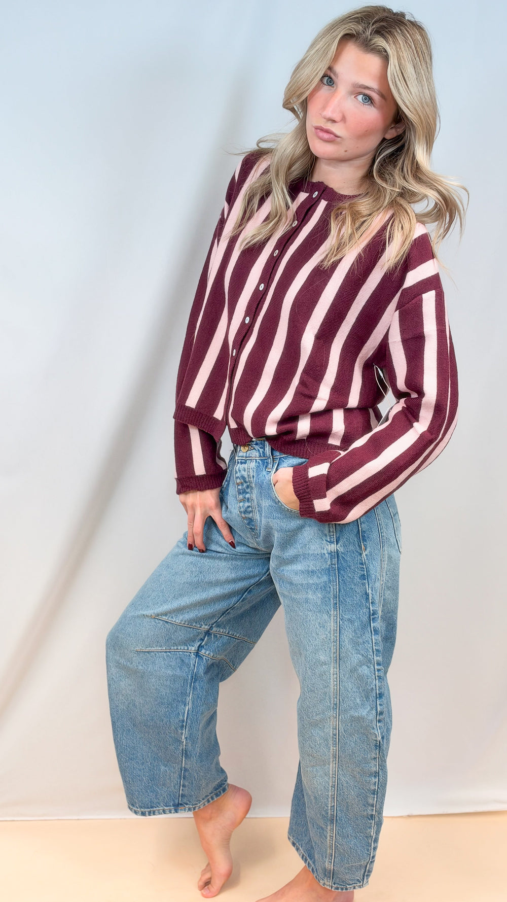 Woman wearing burgundy and pink stripe cardigan with barrel jeans on plain background