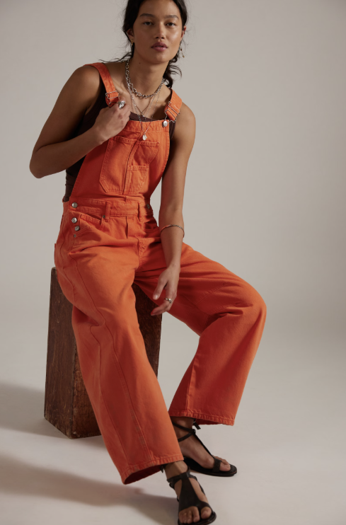 Person wearing orange overalls sitting on a wooden block against a plain background