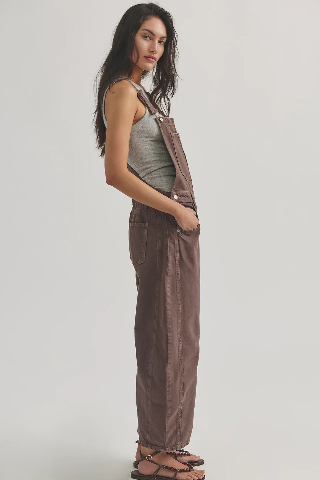 Woman shown from side view wearing brown overalls with grey tank on plain background