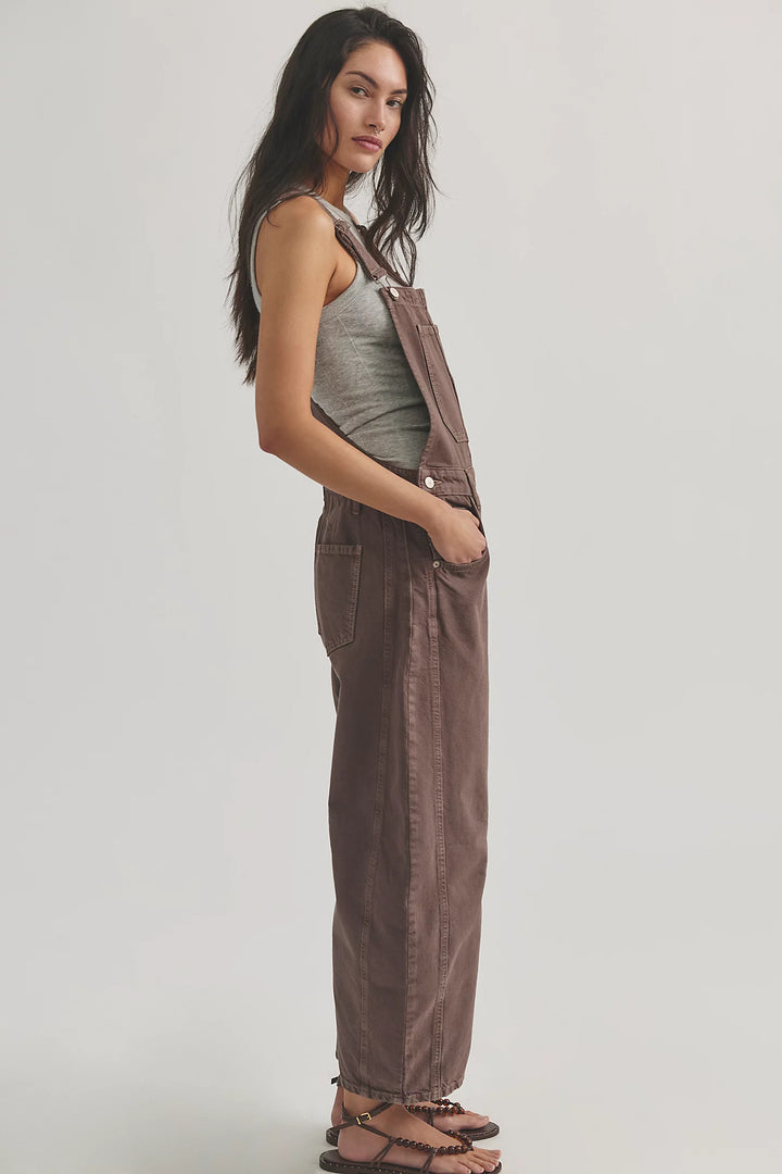 Woman shown from side view wearing brown overalls with grey tank on plain background