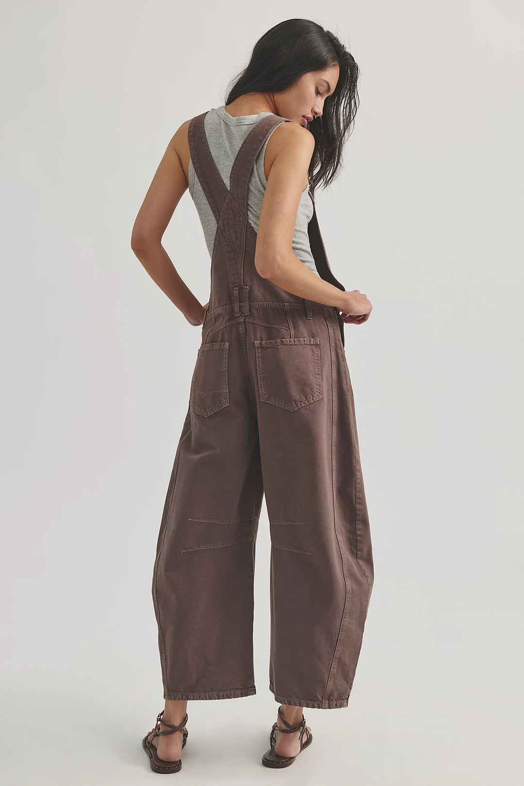 Woman shown from back view wearing brown overalls with grey tank on plain background