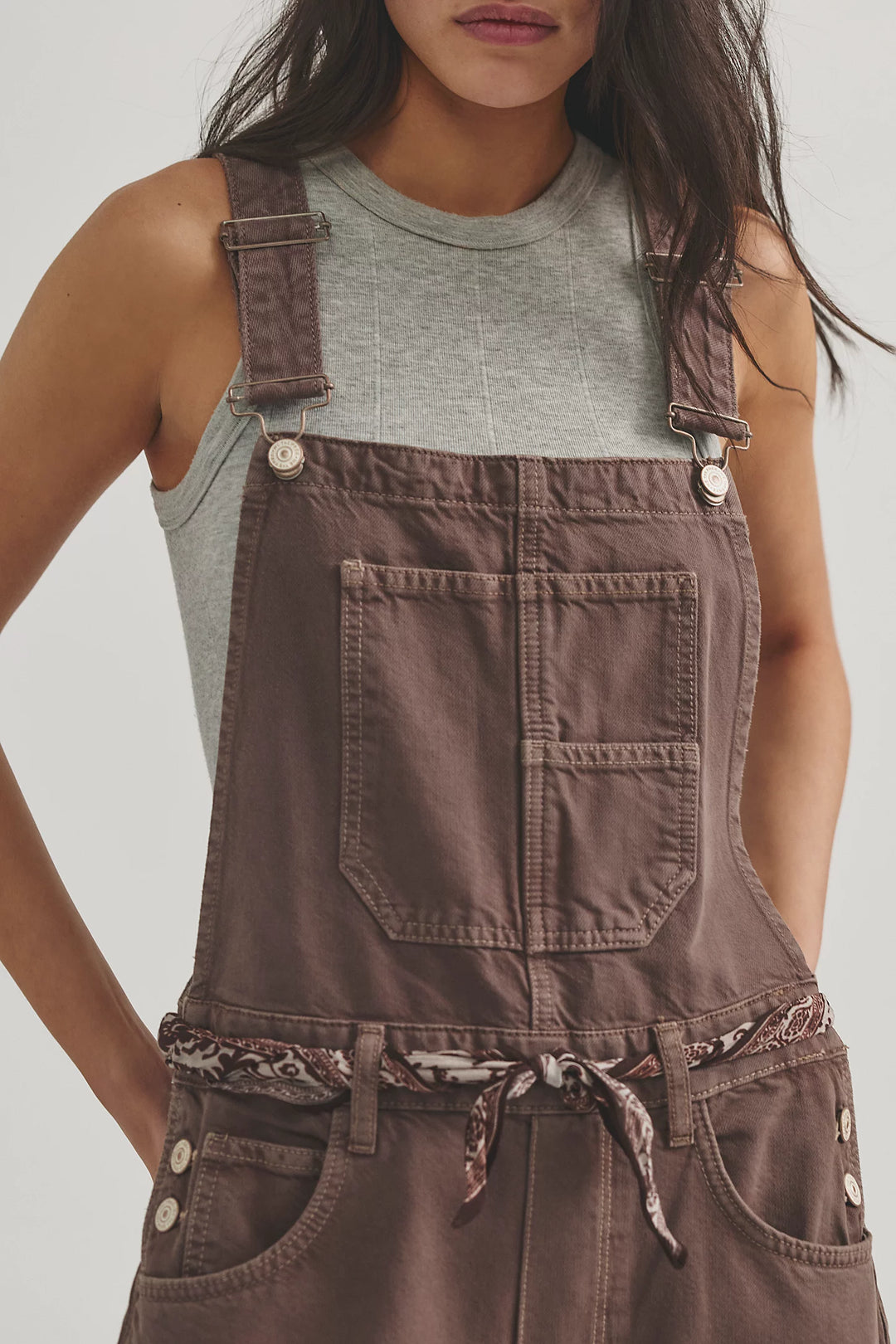 Close up of woman wearing brown overalls with grey tank and scarf belt on plain background