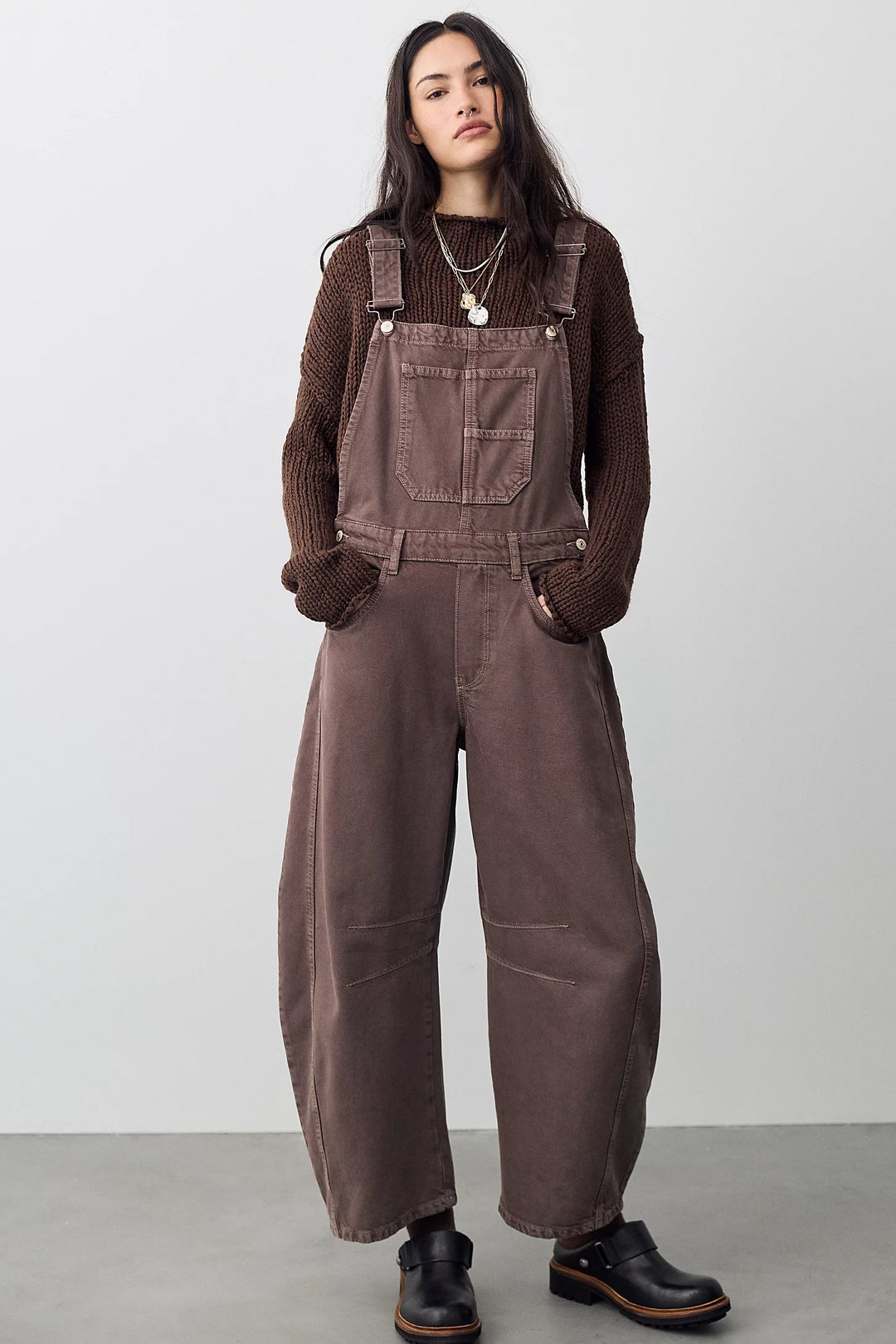 Woman wearing brown barrel overalls with brown sweater on plain background