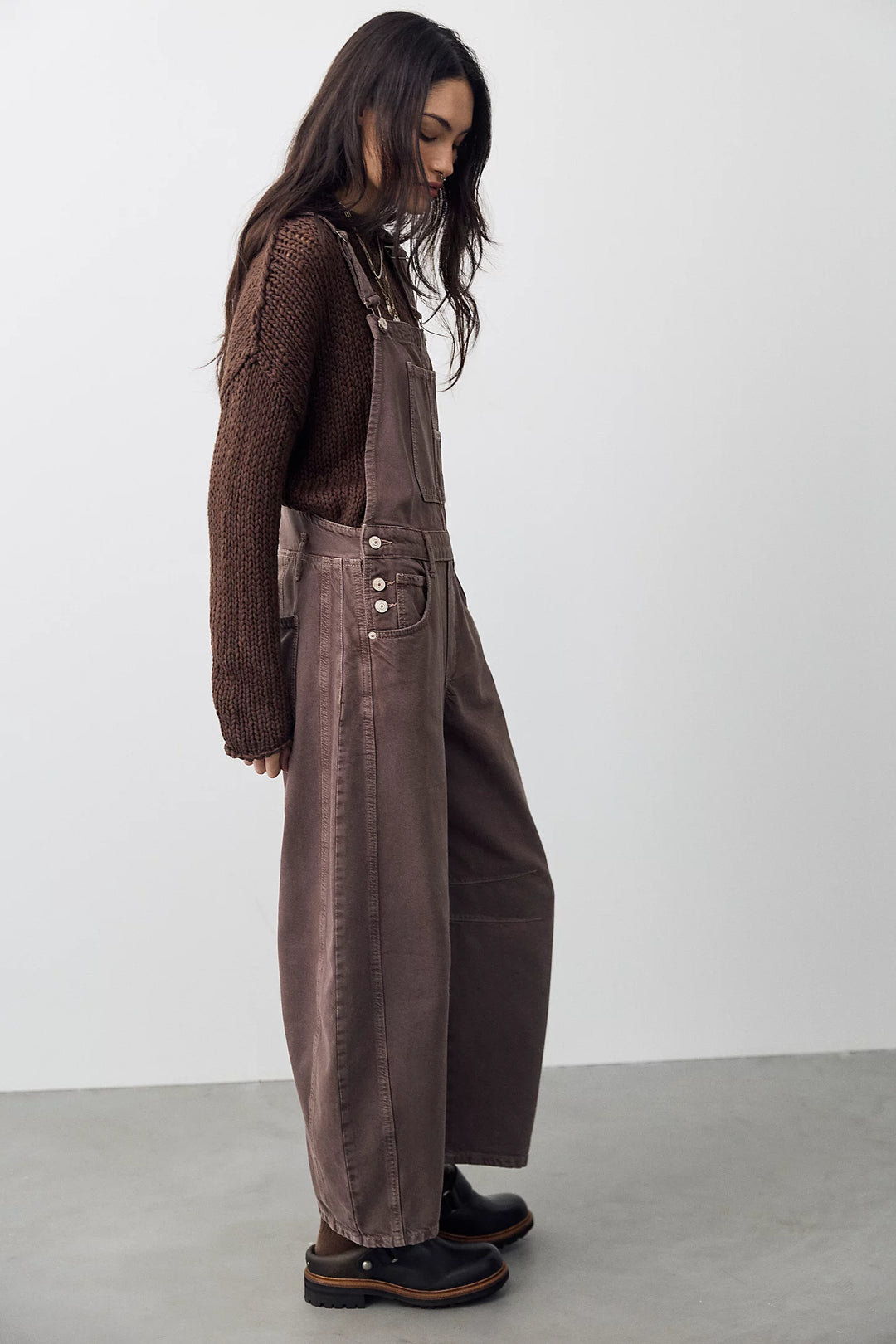 Woman shown from side view wearing brown overalls with brown sweater on plain background