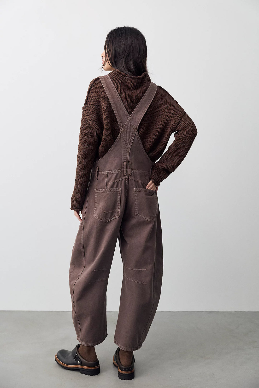 Woman shown from back view wearing brown overalls with brown sweater on plain background