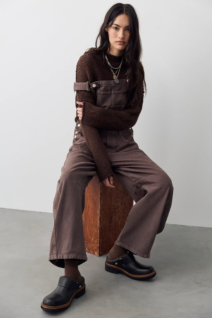 Woman sitting on wooden box wearing brown overalls with brown sweater on plain background