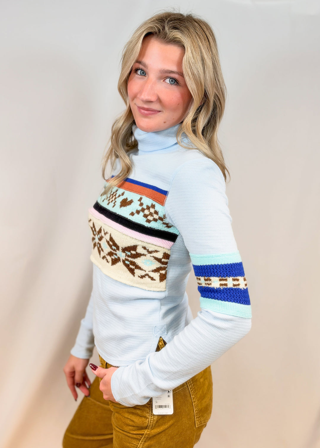 Woman shown from side wearing light blue turtle neck top with argyle and snowflake designs with camel cord pants on plain background